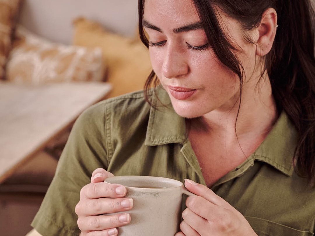 Woman in a green shirt sitting indoors, holding a mug with both hands and looking down thoughtfully.